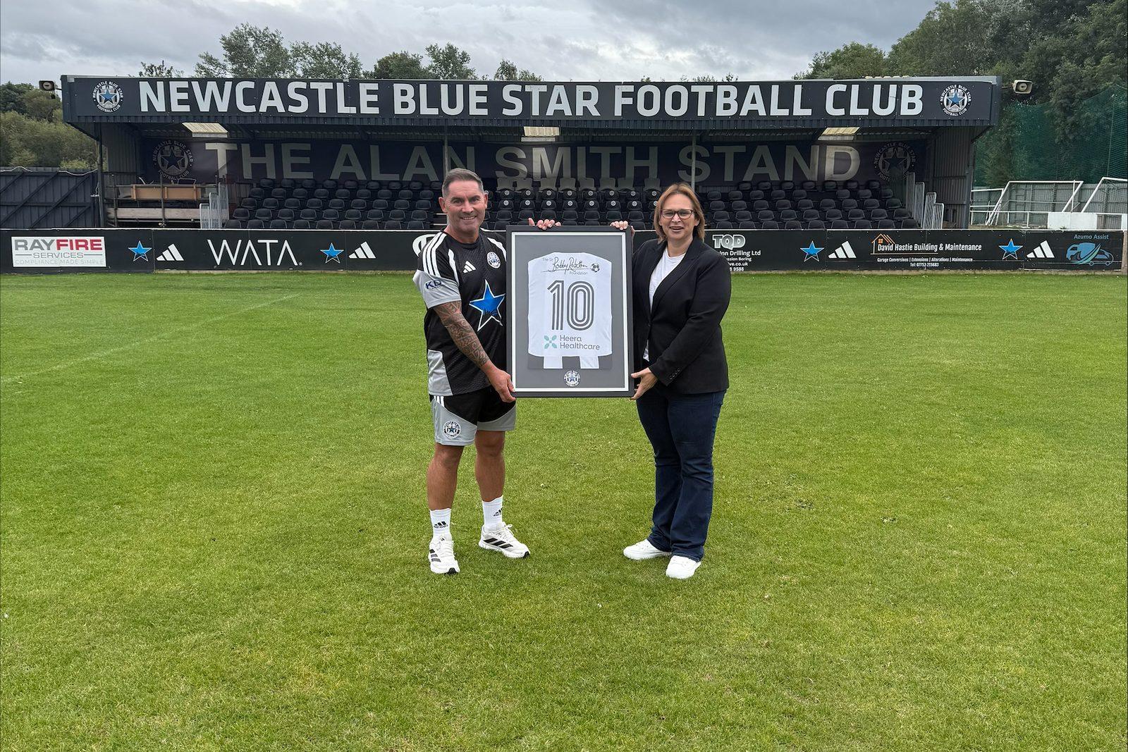 Representatives of Heera Healthcare and Newcastle Blue Star FC holding a framed number 10 shirt on the pitch in front of the Alan Smith Stand